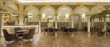 A spacious indoor area with a high ceiling, featuring a central seating area with round tables and chairs, surrounded by arches with decorative tile work.