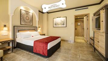 A hotel room with a large bed, a red throw, a framed picture on the wall, and a sign above the bed that says "Somos Pet Friendly".