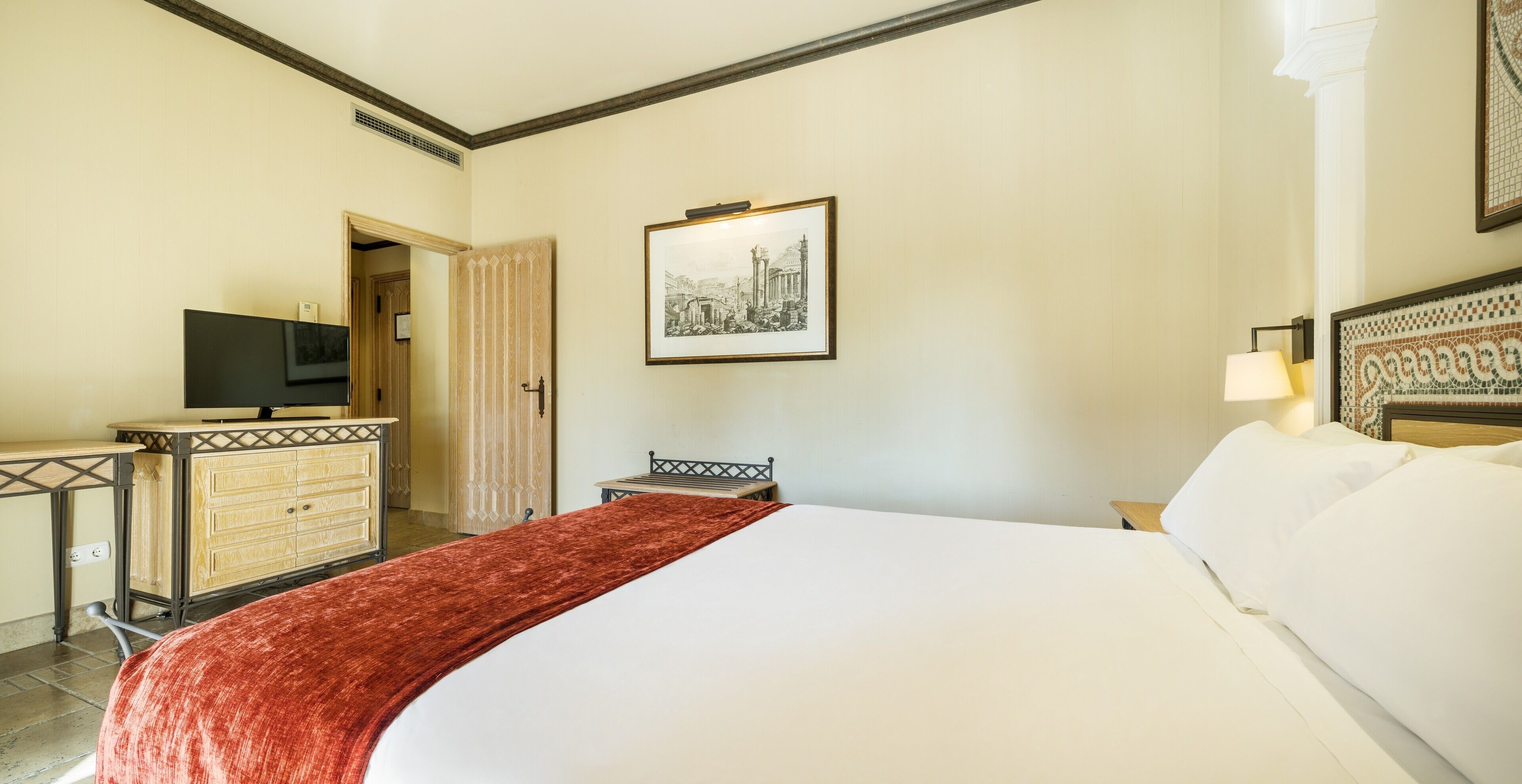 A hotel room with a bed, a desk with a television, a chair, and a framed picture on the wall.