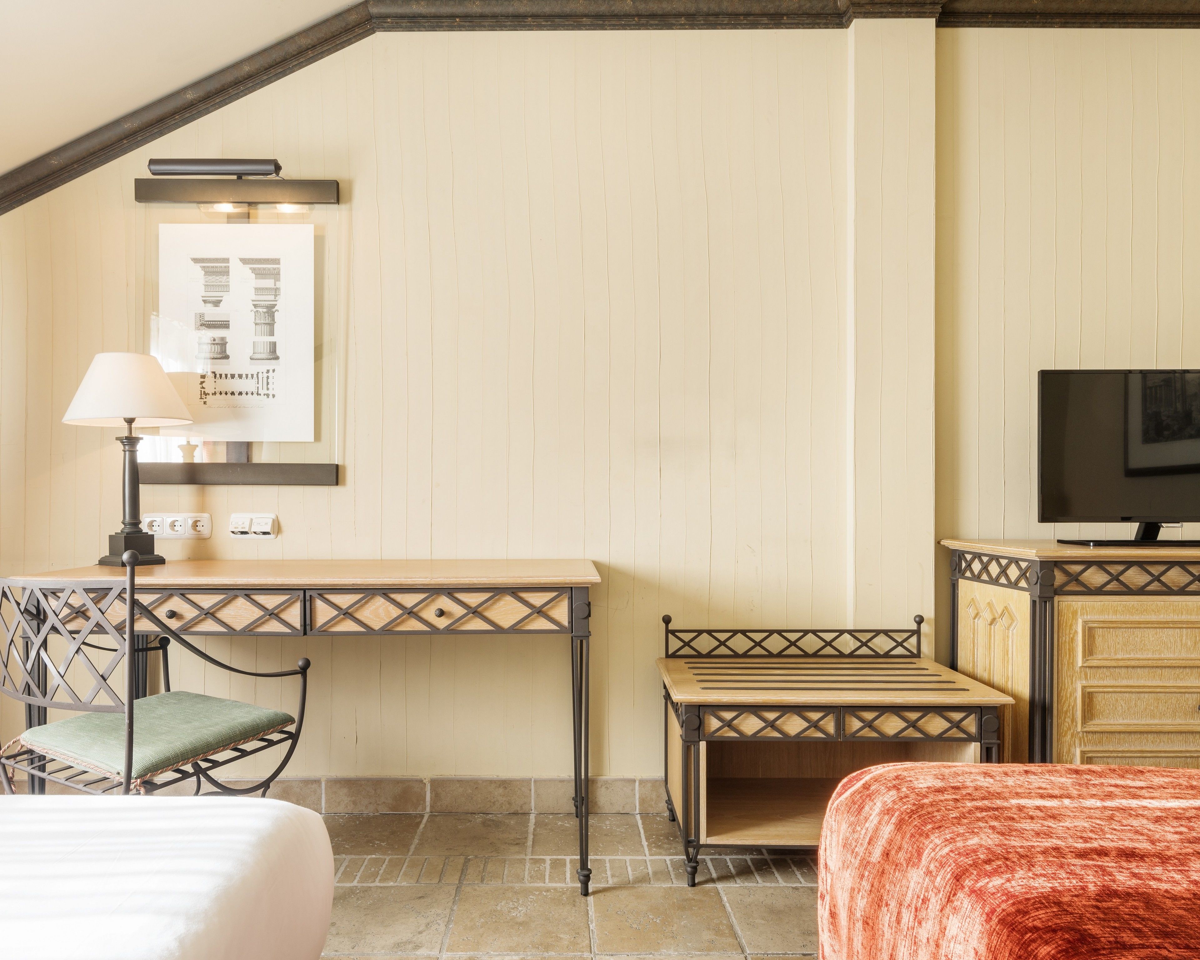 Habitación de hotel con un escritorio de madera, una silla, una cama con manta naranja, un televisor y un cuadro en la pared.