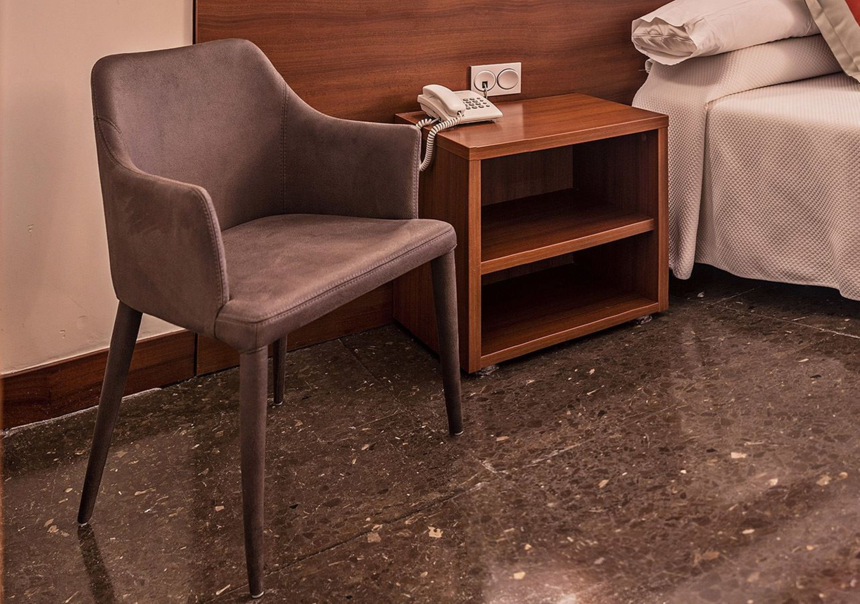 A modern hotel room with a grey upholstered armchair, a wooden nightstand with a telephone, and a bed with a white cover.