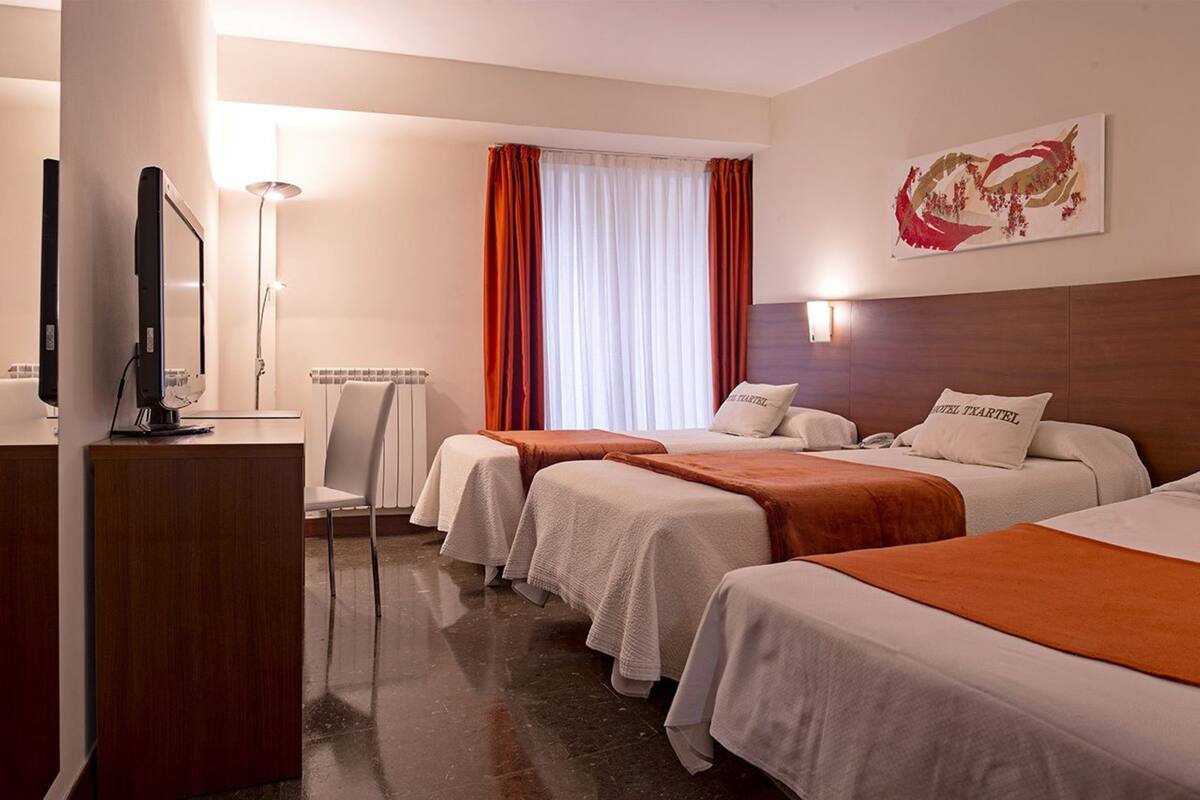 A hotel room with two beds, a desk, a chair, a TV, and a window with curtains.
