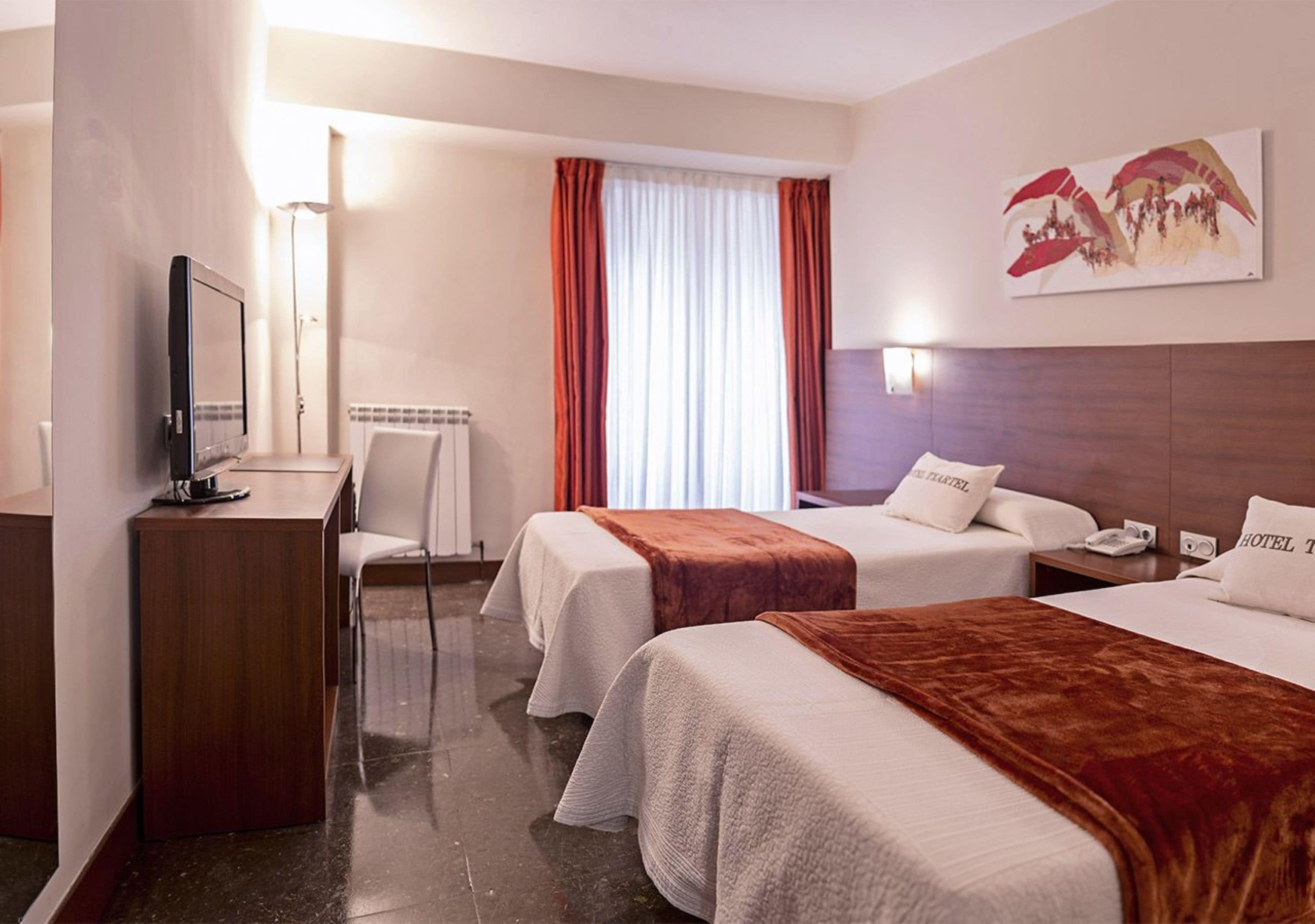 Habitación de hotel con dos camas, un escritorio, una silla, un televisor y una ventana con cortinas.