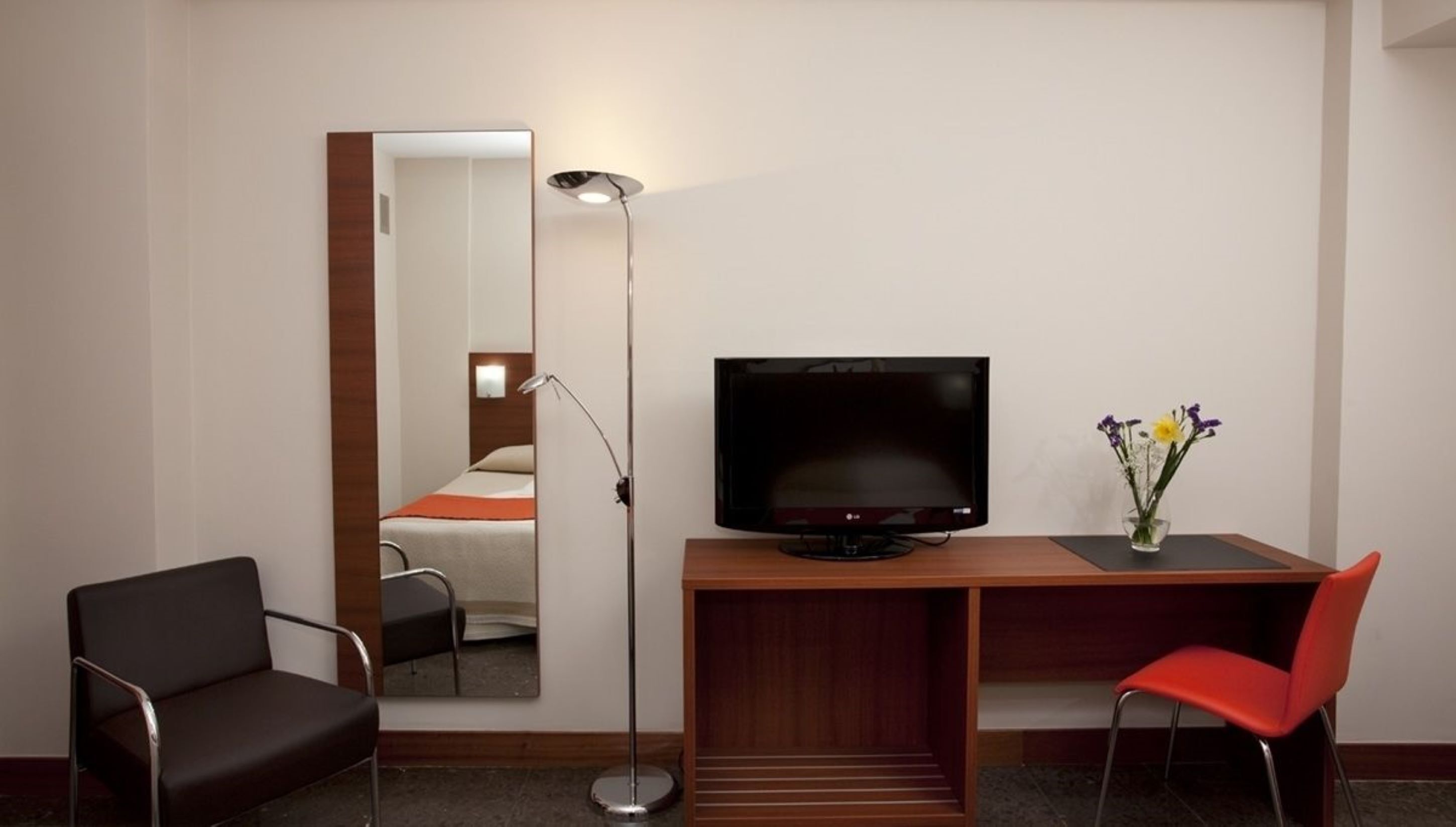 A hotel room with a bed, a desk with a TV, a chair, a floor lamp, and a vase with flowers.