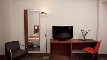 A hotel room with a bed, a desk with a TV, a chair, a floor lamp, and a vase with flowers.