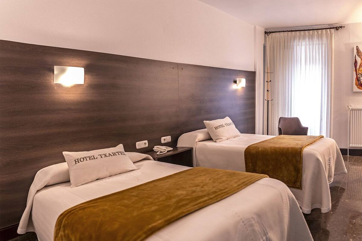 A hotel room with two beds, each with a pillow labeled "HOTEL TXARTEN." There are wall-mounted light fixtures and a chair in the corner.