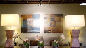 Um quarto com duas pinturas abstratas na parede, uma mesa de madeira com luminárias e vasos de flores, e teto com vigas aparentes.