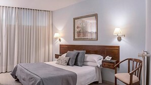 Quarto de hotel com cama, cadeira, mesa de cabeceira e luminária fixada na parede.