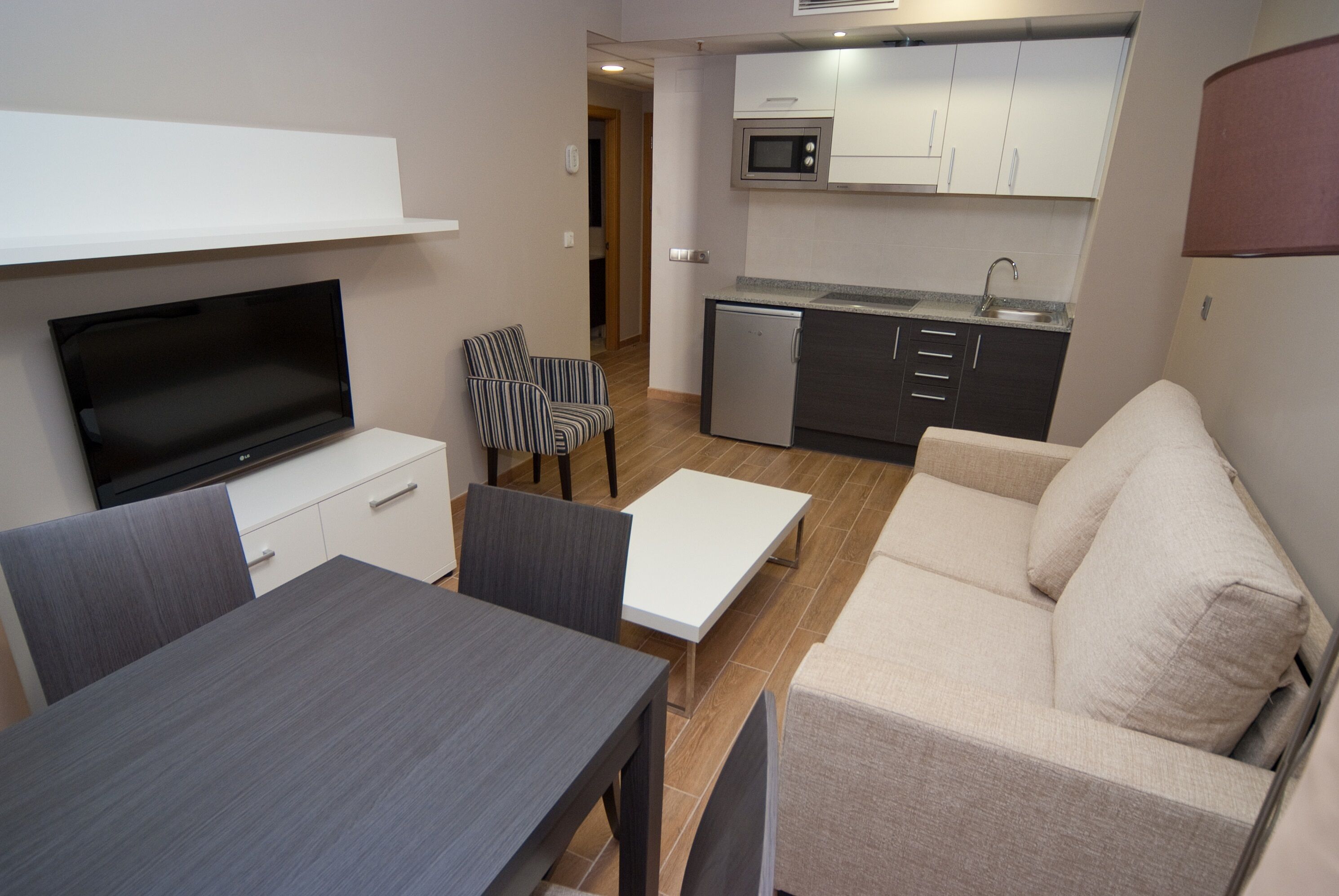 A compact living space with a flat-screen TV, a dining table, a sofa, a kitchenette, and striped chairs.