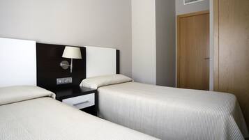 A hotel room with two single beds, a bedside table with a lamp, a power outlet, and a wooden door.