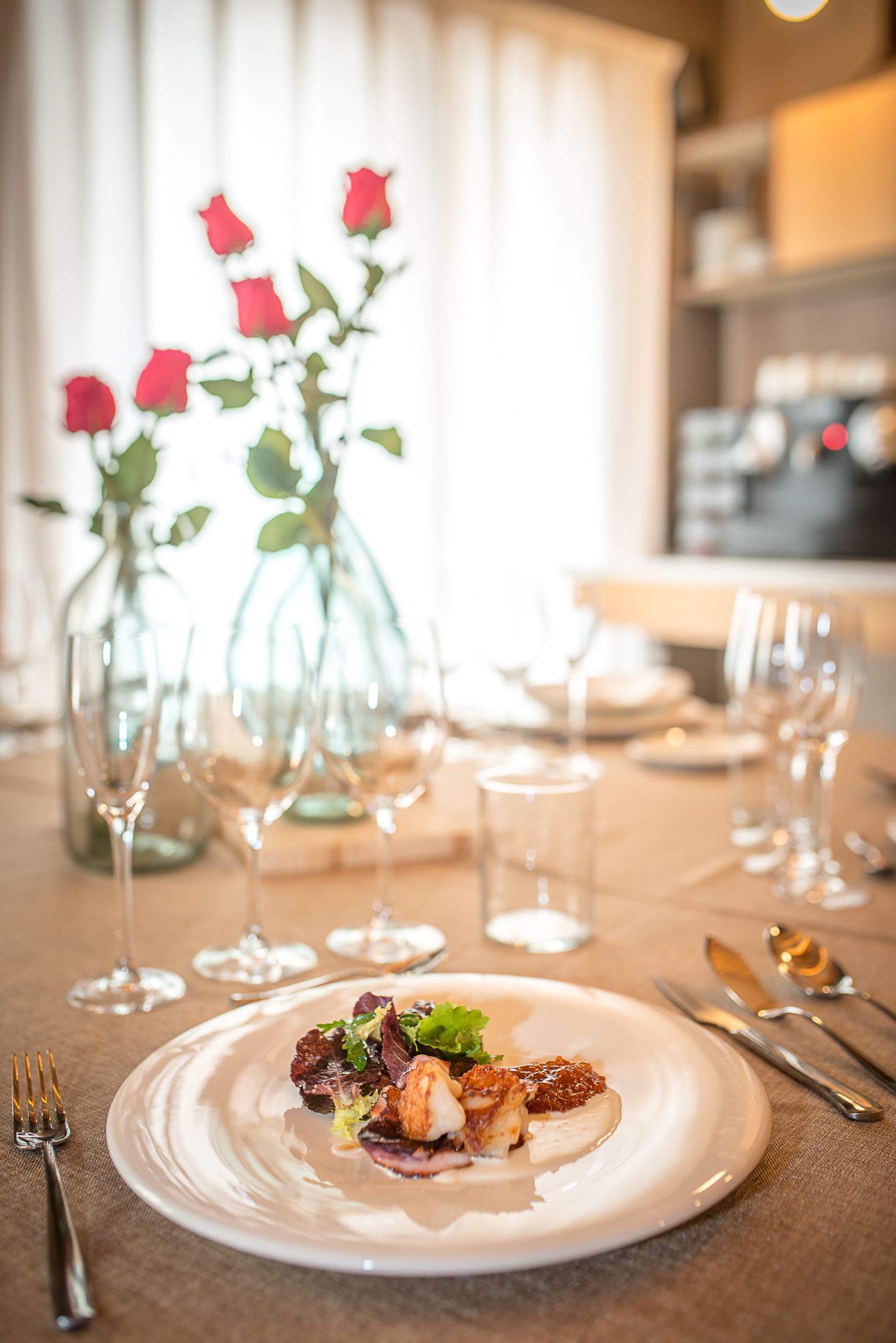 A dining table set with plates, cutlery, and wine glasses, with a vase of red roses in the background.