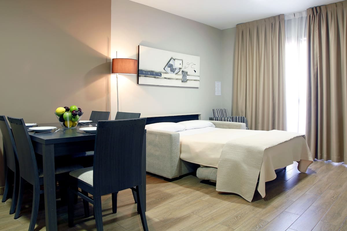 A hotel room with a bed, a desk with a lamp, a chair, and a dining table with a fruit bowl.