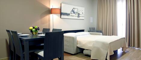 A hotel room with a bed, a desk with a lamp, a chair, and a dining table with a fruit bowl.