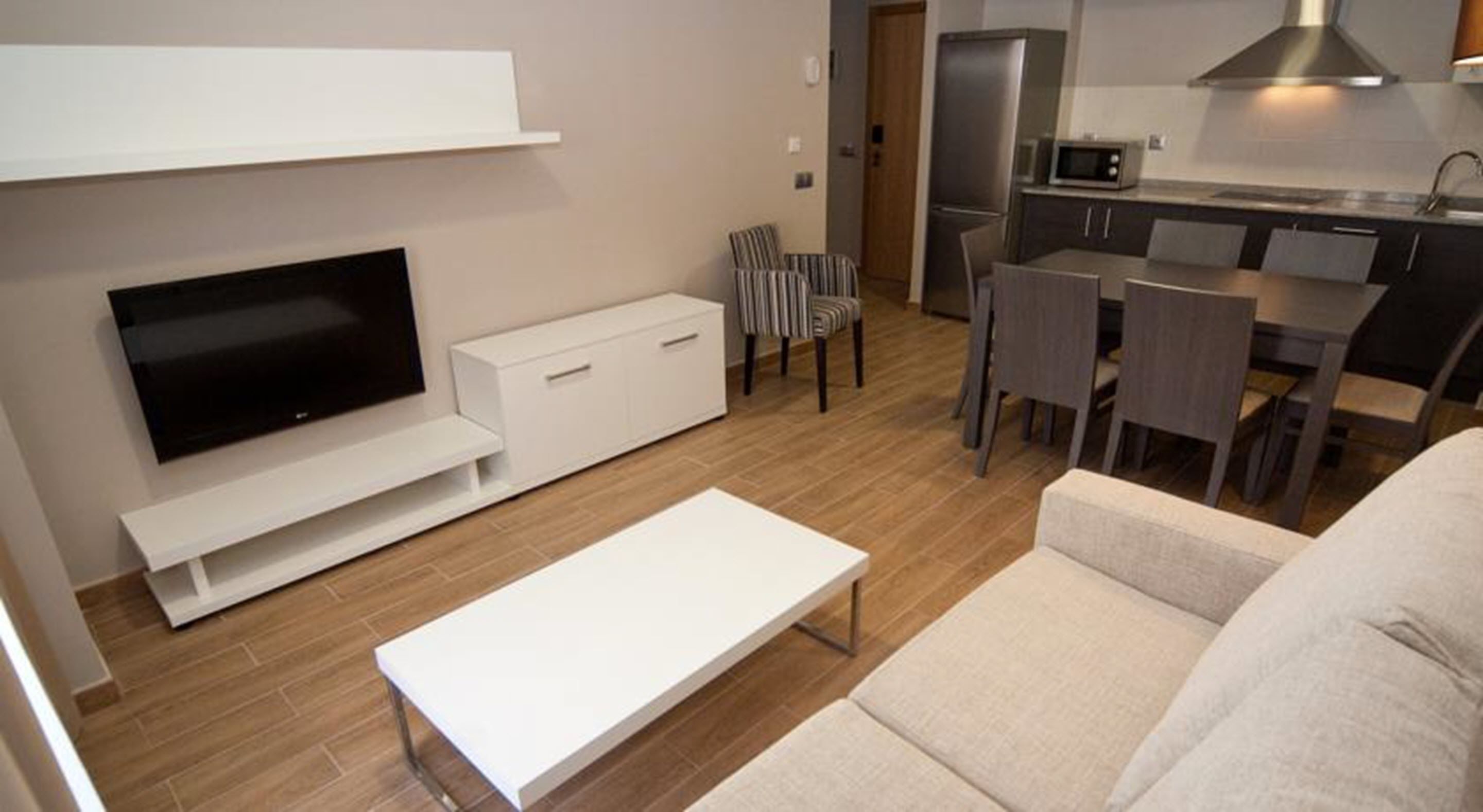 Una sala de estar moderna con una televisión de pantalla plana, un sofá blanco, una mesa de centro y un comedor con una mesa y sillas.