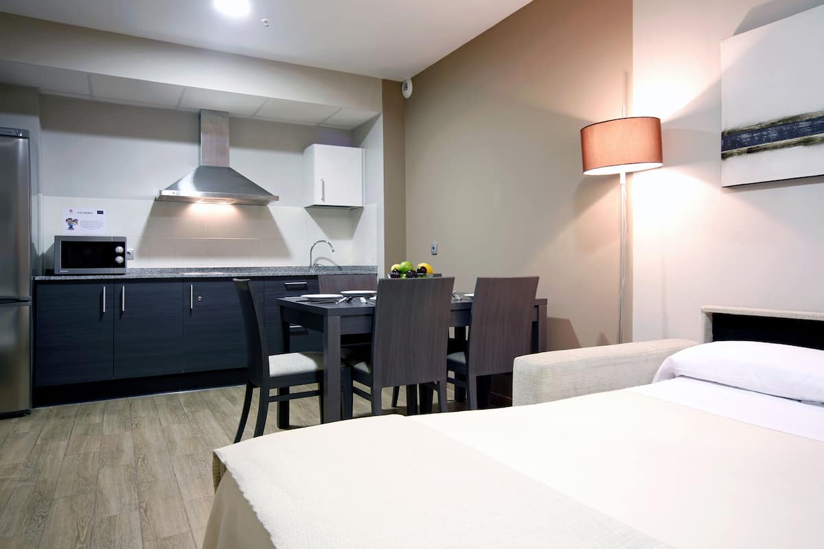 A hotel room with a bed, a dining table with chairs, a kitchen area with a microwave, and a lamp.