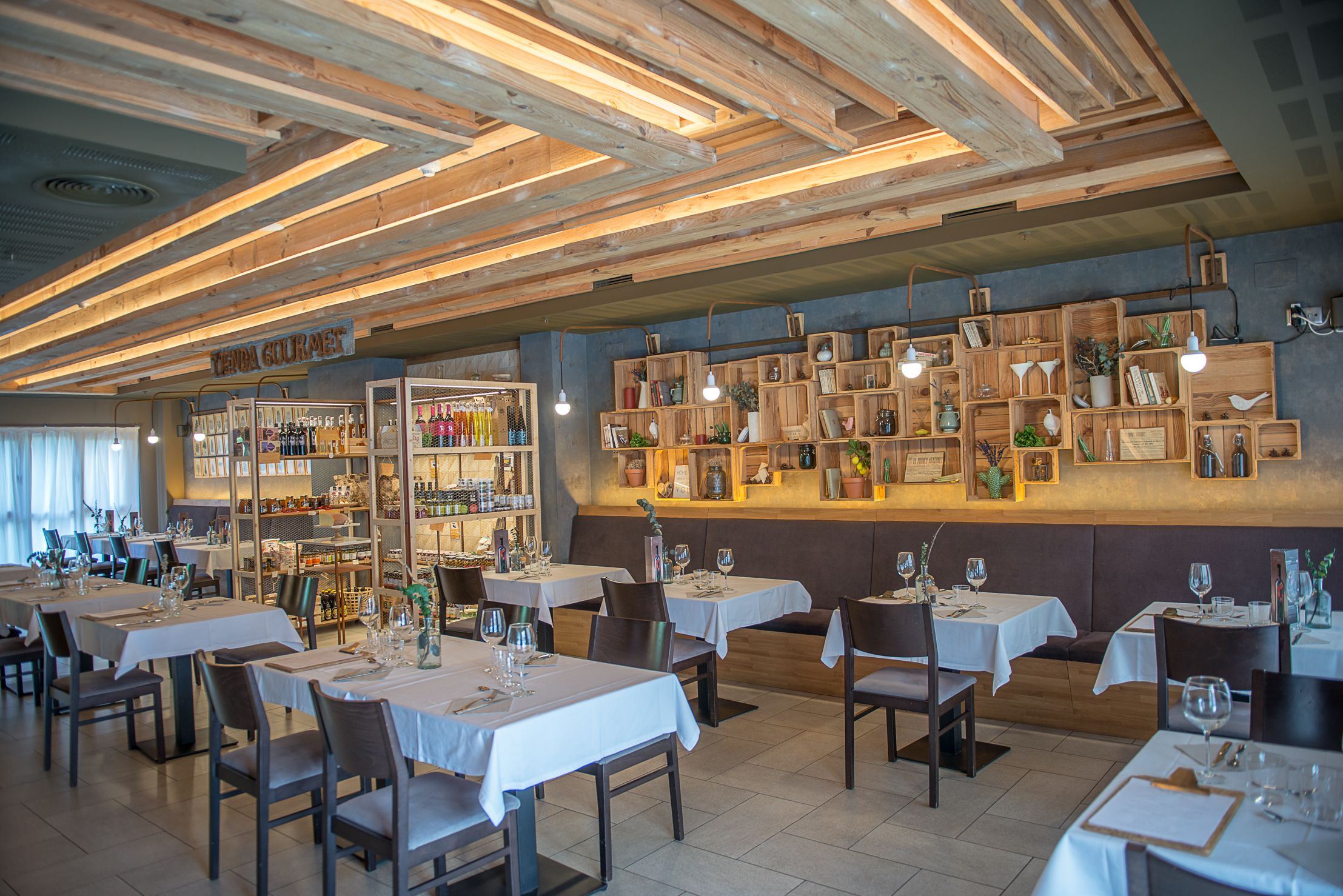 Interior de restaurante con vigas de madera en el techo, manteles blancos y estantes con diversos objetos.