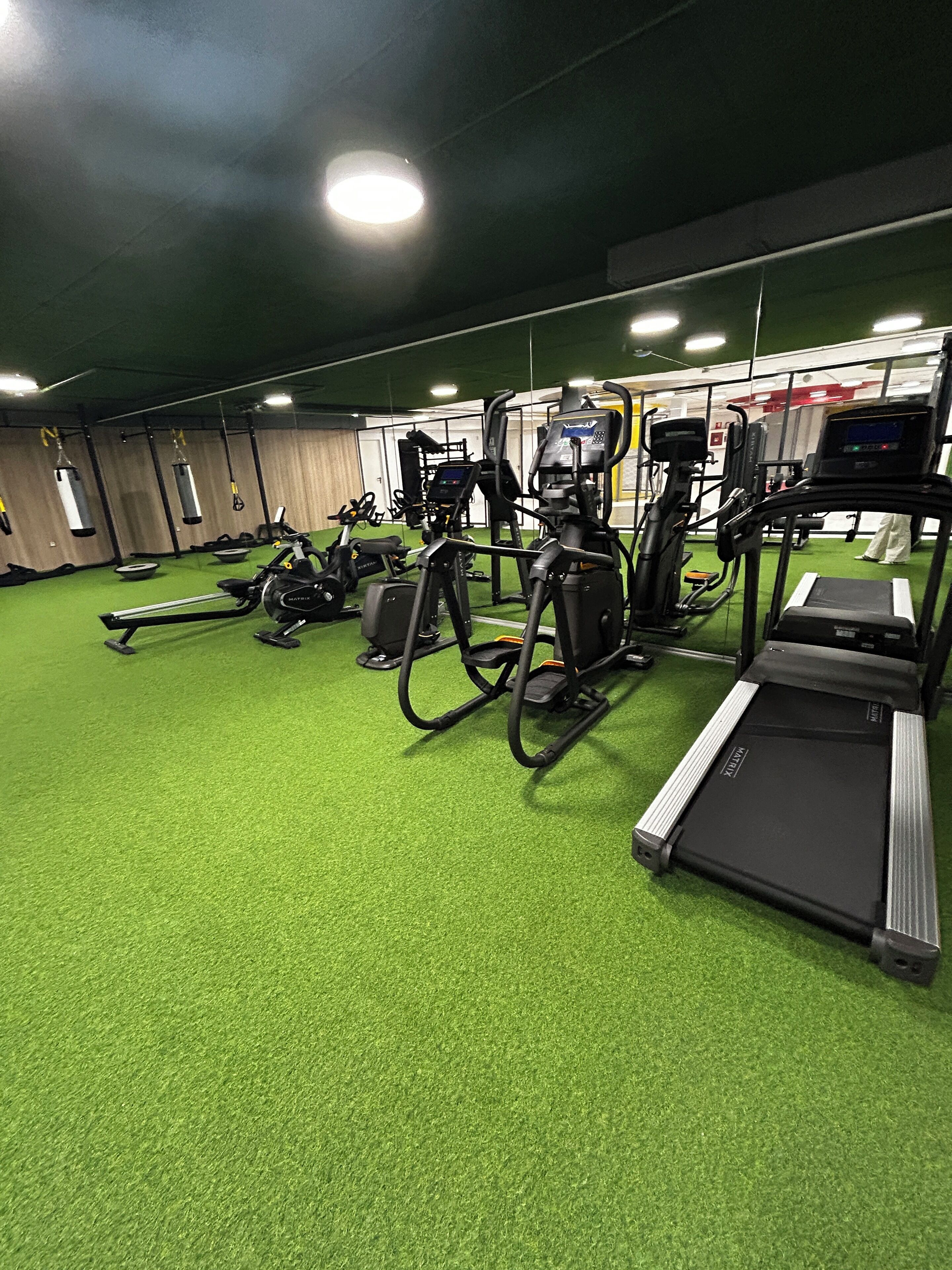 A gym with various exercise equipment, including treadmills, stationary bikes, and weight machines, on a green artificial turf floor.