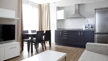 Eine moderne Küche mit dunklen Schränken, einem Edelstahl-Kühlschrank und einer Mikrowelle. Ein Essbereich mit weißem Tisch und Stühlen, sowie ein Flachbildfernseher auf einem weißen Schrank.