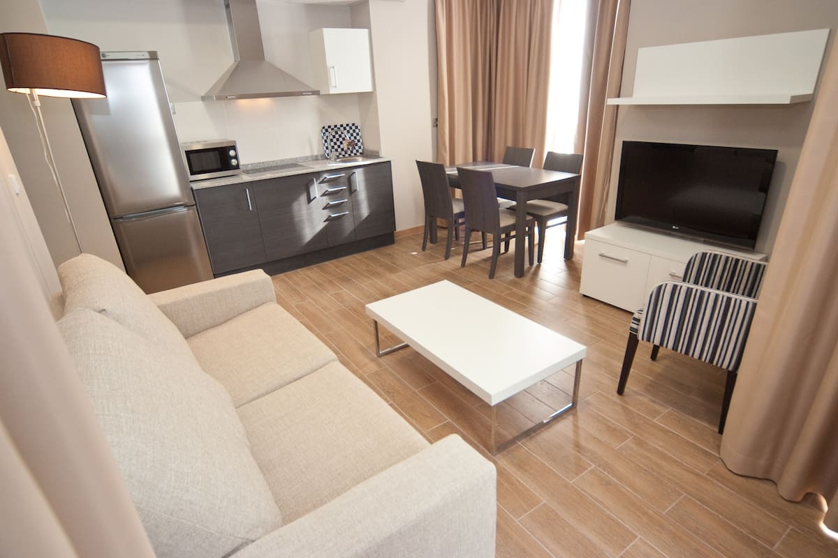 A modern living room with a sofa, a coffee table, a TV, a dining area with chairs, and a kitchen with a microwave and refrigerator.