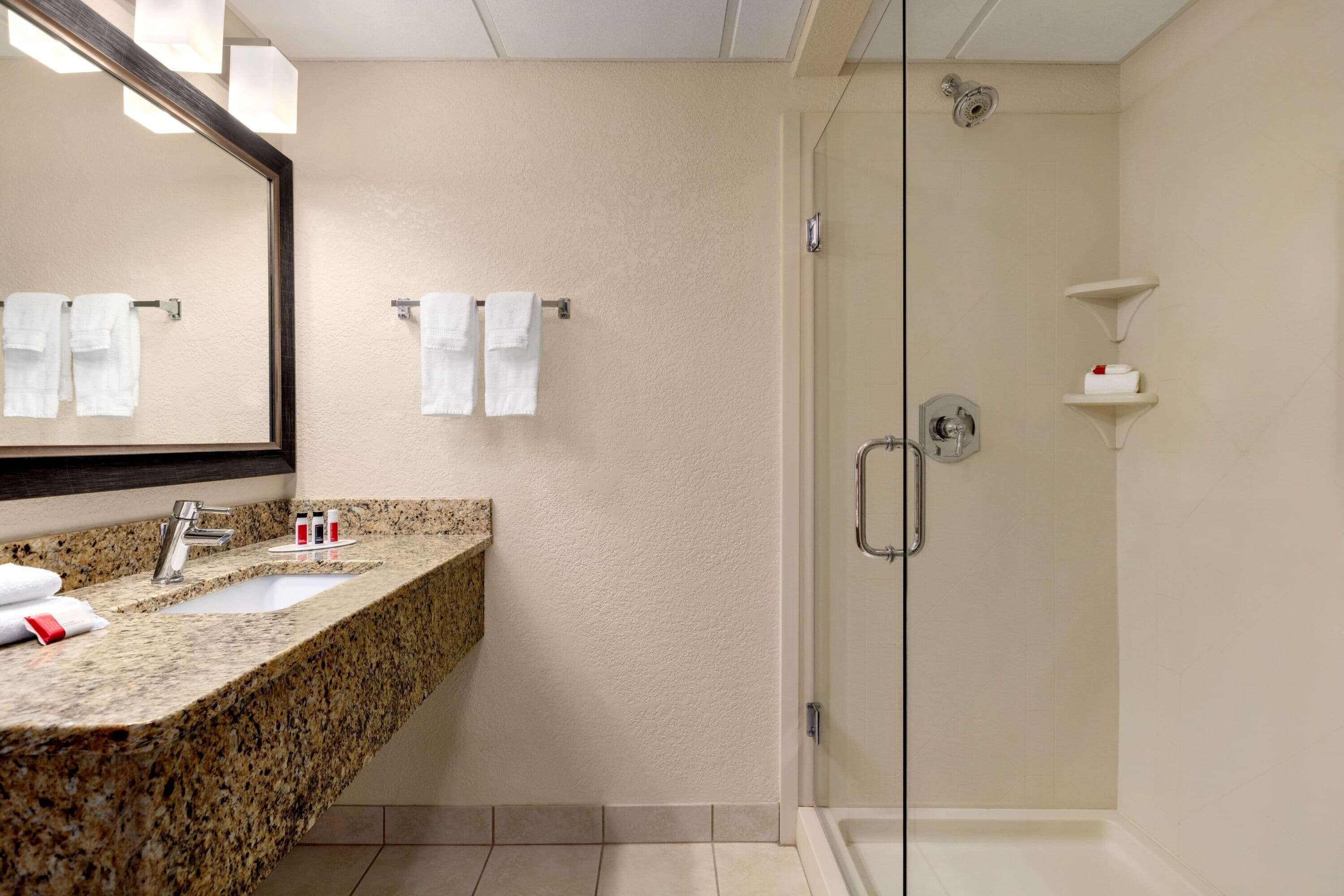 A modern bathroom with a granite countertop, a large mirror, two towel racks, a glass-enclosed shower, and a white sink.