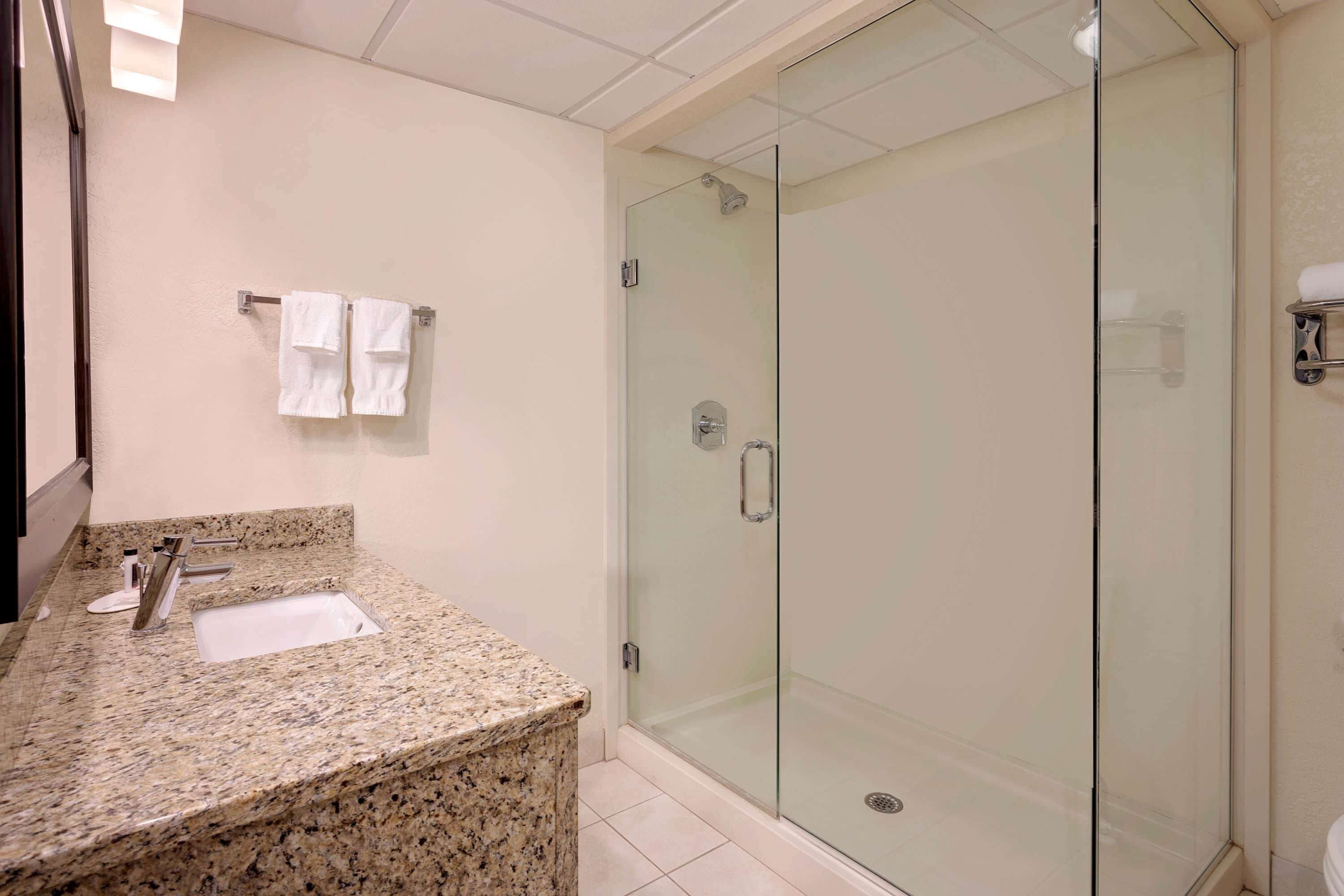 A modern bathroom with a granite countertop, a glass-enclosed shower, and white towels hanging on a rack.
