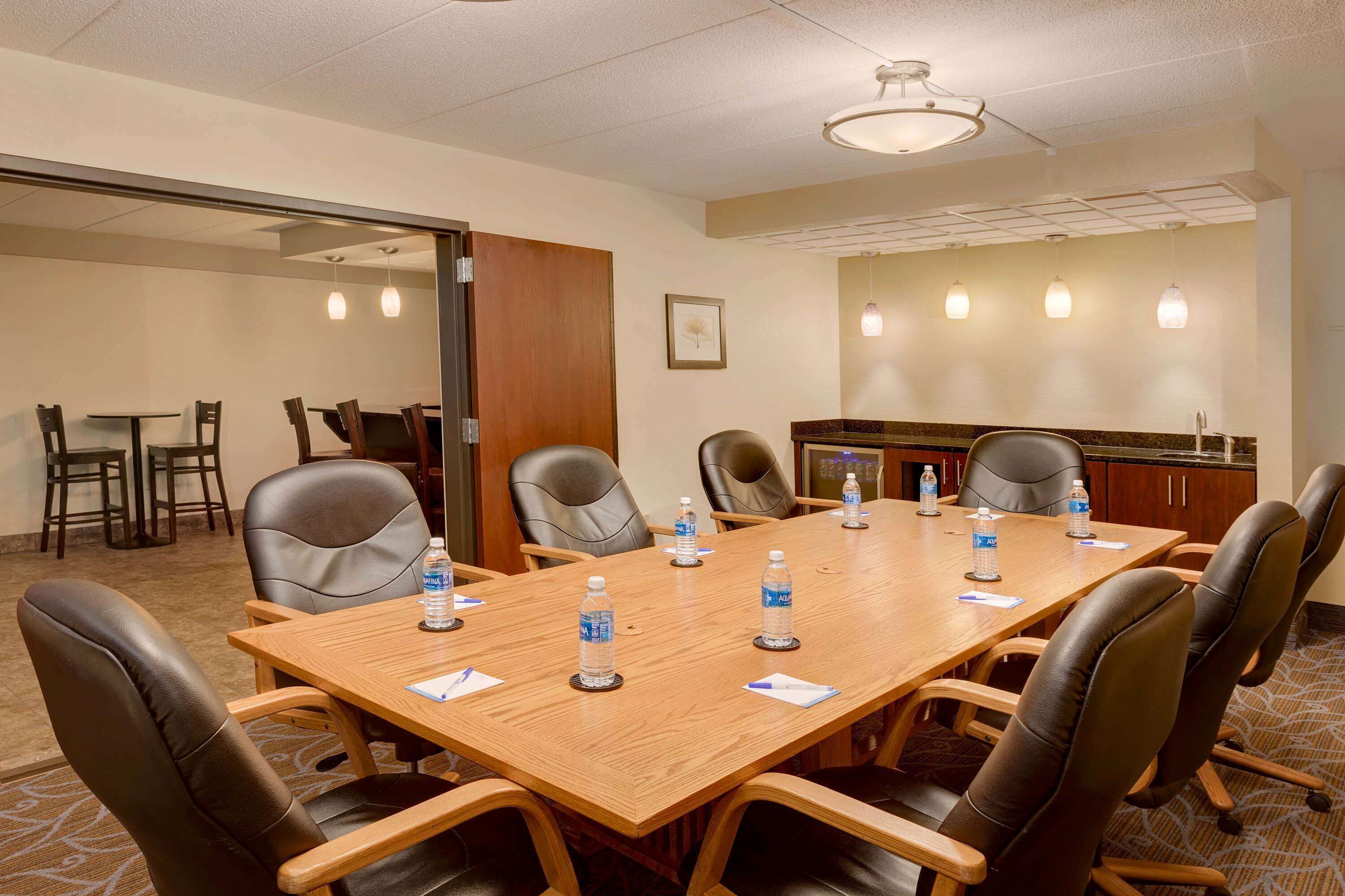 A conference room with a long wooden table, black leather chairs, water bottles, and a kitchen area in the background.