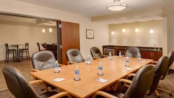 A conference room with a long wooden table, black leather chairs, water bottles, and a kitchen area in the background.