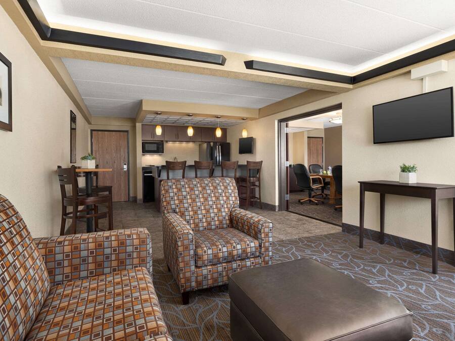 Un vestíbulo de hotel con un sofá y un sillón estampados, un televisor de pantalla plana, una pequeña mesa con una planta y una zona de cocina al fondo.