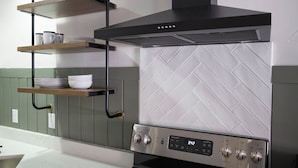 A kitchen with a black range hood, stainless steel cooktop, and a digital display. There are wooden shelves with dishes and a white tiled backsplash. - River Canyon Lodge (Moab)