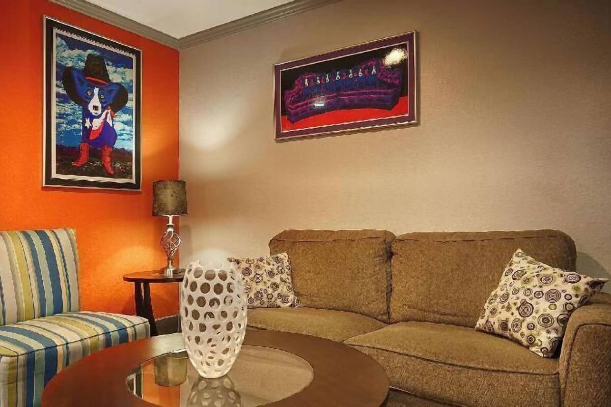 A living room with a striped armchair, a brown sofa with patterned cushions, a glass coffee table, and two framed artworks on the walls.