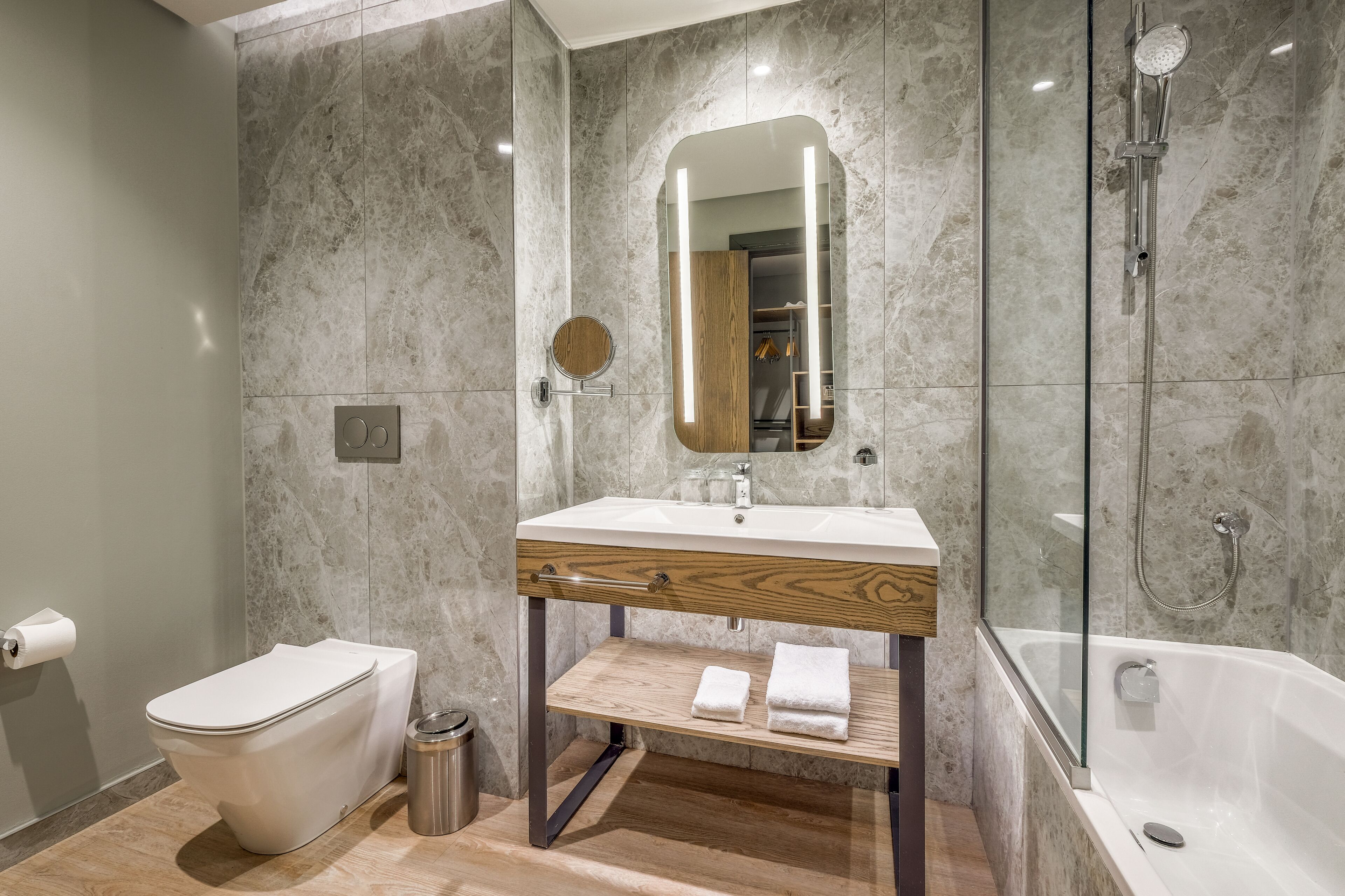 A modern bathroom with a marble wall, a wooden vanity, a toilet, a glass shower enclosure, and a bathtub.
