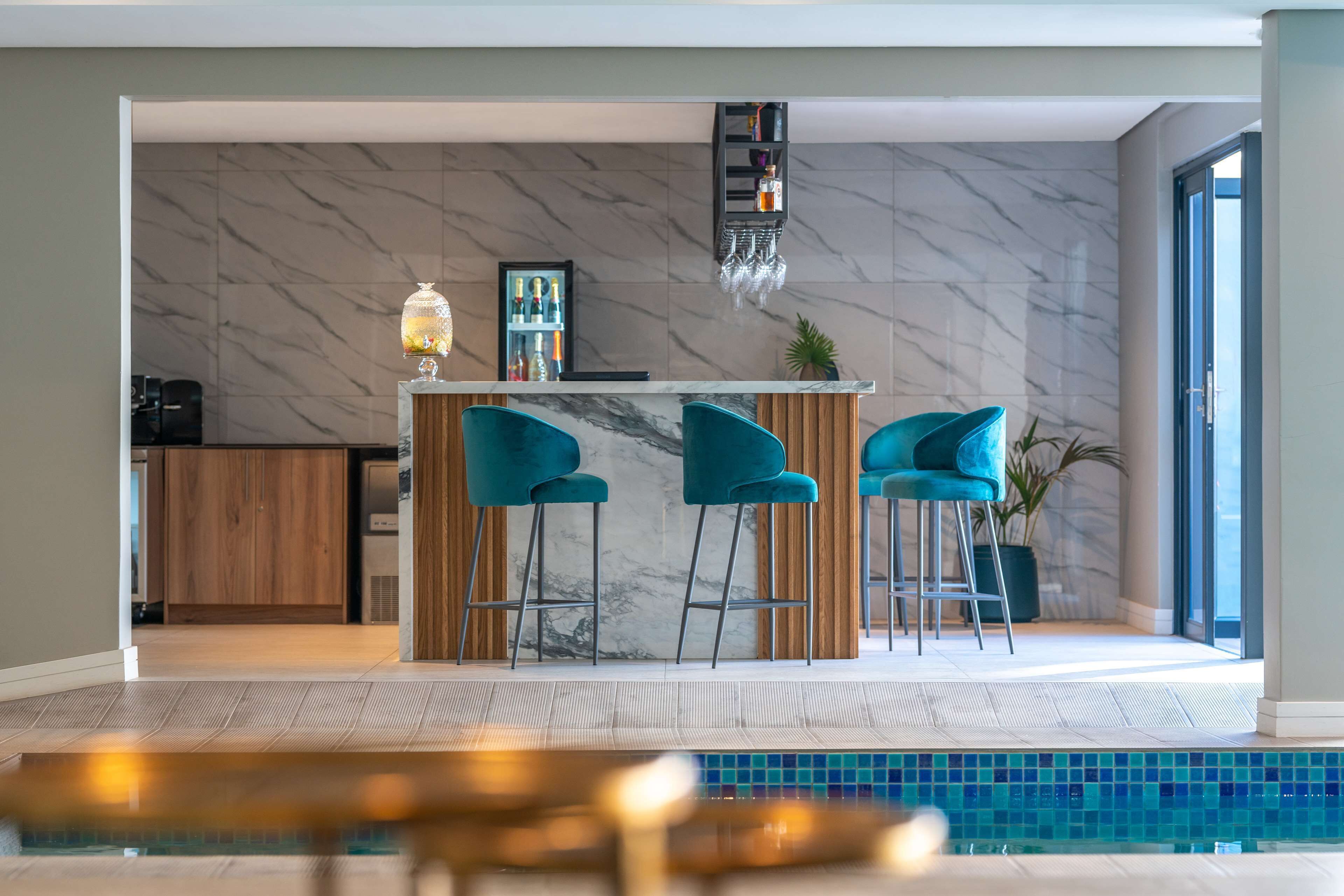 A modern kitchen with marble walls, a bar area with teal chairs, and a pool area with a tiled floor.