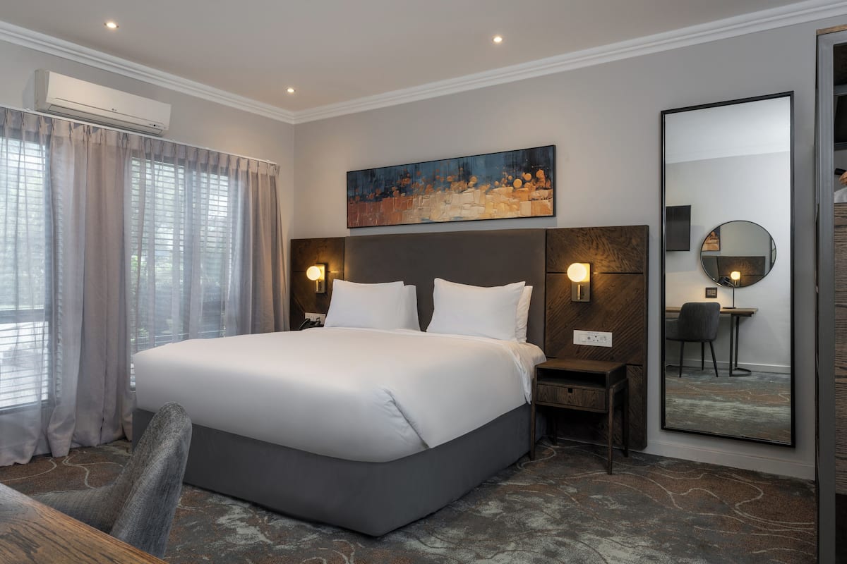 A modern hotel room with a large bed, a headboard with integrated lighting, a painting above the bed, a large mirror, and a desk with a chair.