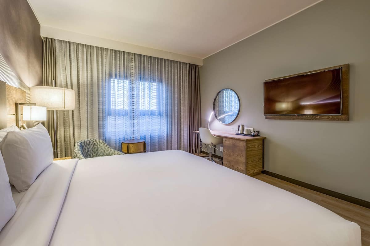 A hotel room with a large bed, a desk with a chair, a round mirror, and a wall-mounted TV.