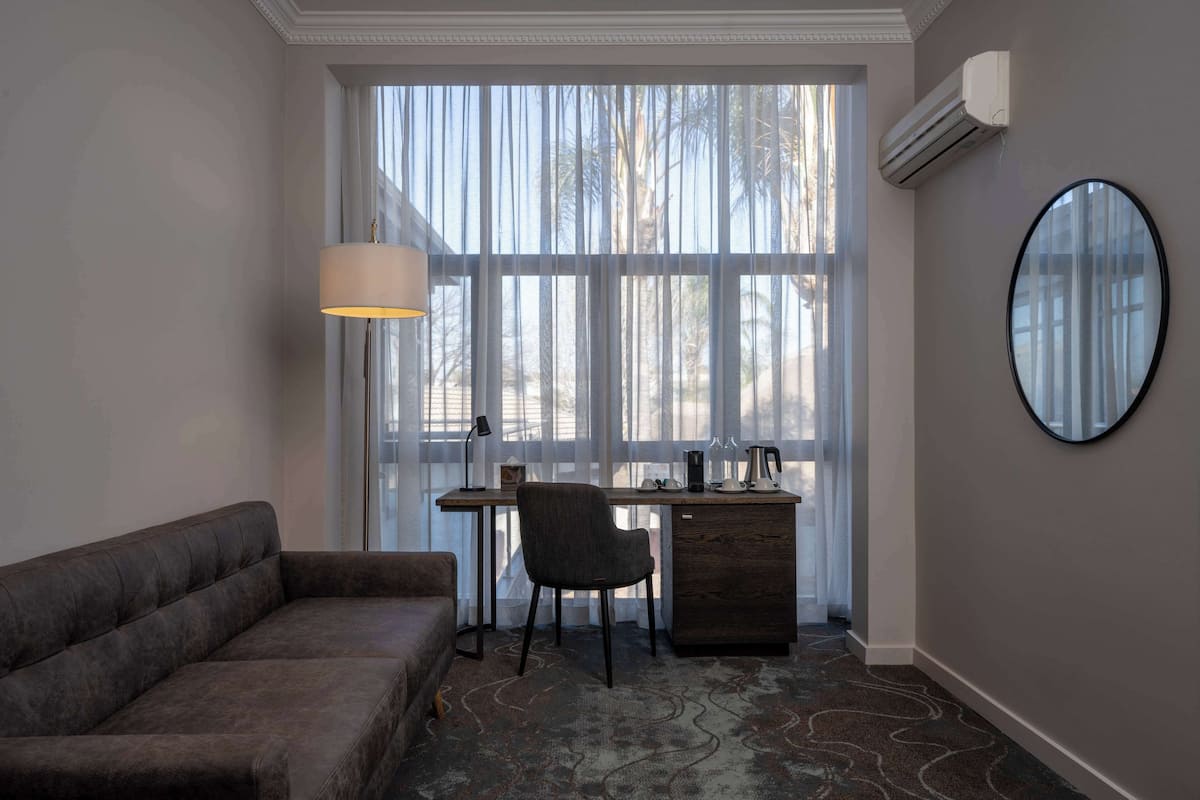 A room with a sofa, a desk with a chair, a large window with sheer curtains, an air conditioning unit, and a round mirror on the wall.