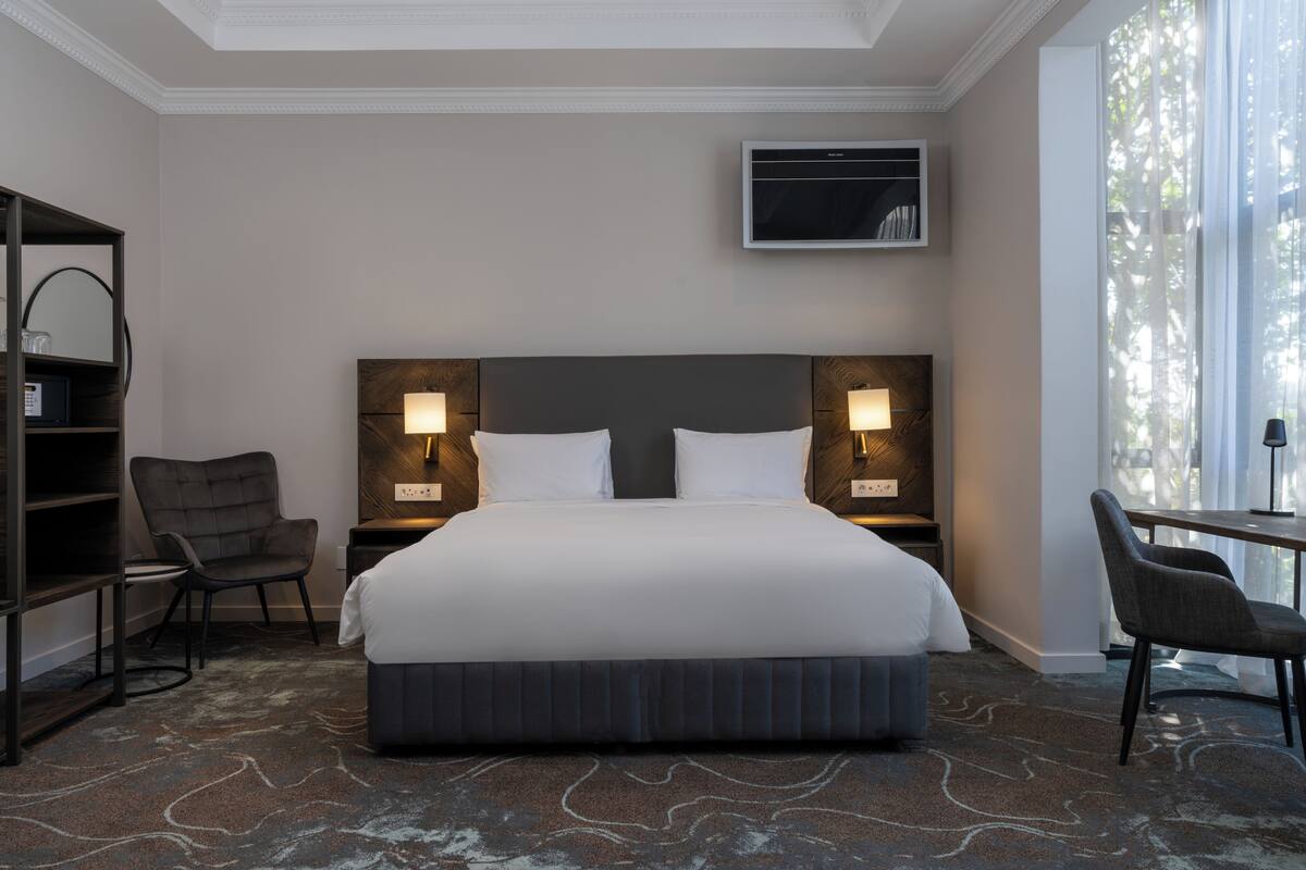 A hotel room with a large bed, two bedside lamps, a TV mounted on the wall, a desk with a chair, and a bookshelf.