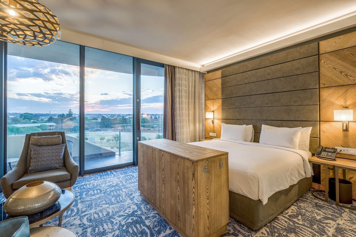 A modern hotel room with a large bed, a wooden nightstand, a desk, a chair, and a view of the city through large windows.