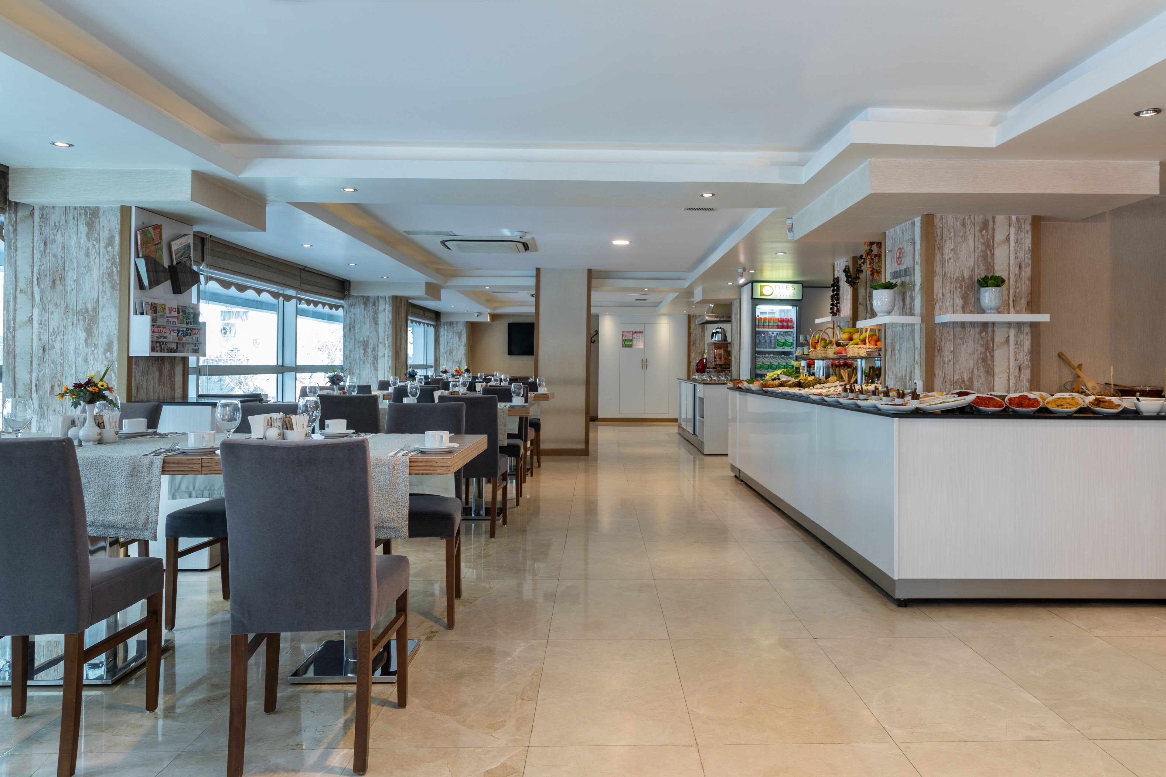 Un comedor de hotel moderno con varias mesas y sillas, una estación de buffet y una cocina al fondo.
