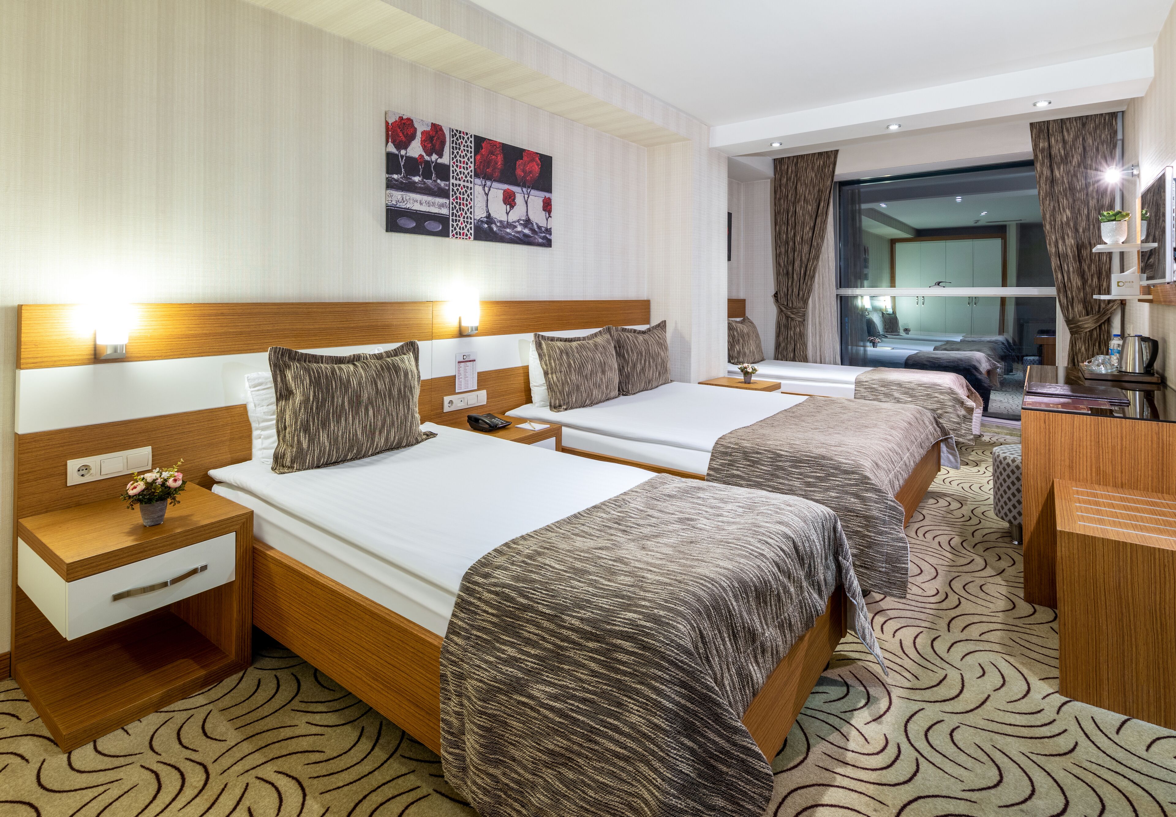 A hotel room with two beds, a wooden headboard, a nightstand with a lamp, a small table with a vase of flowers, and a large mirror.