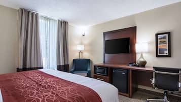 A hotel room with a bed, a desk with a television, a chair, a desk lamp, a microwave, and a refrigerator.