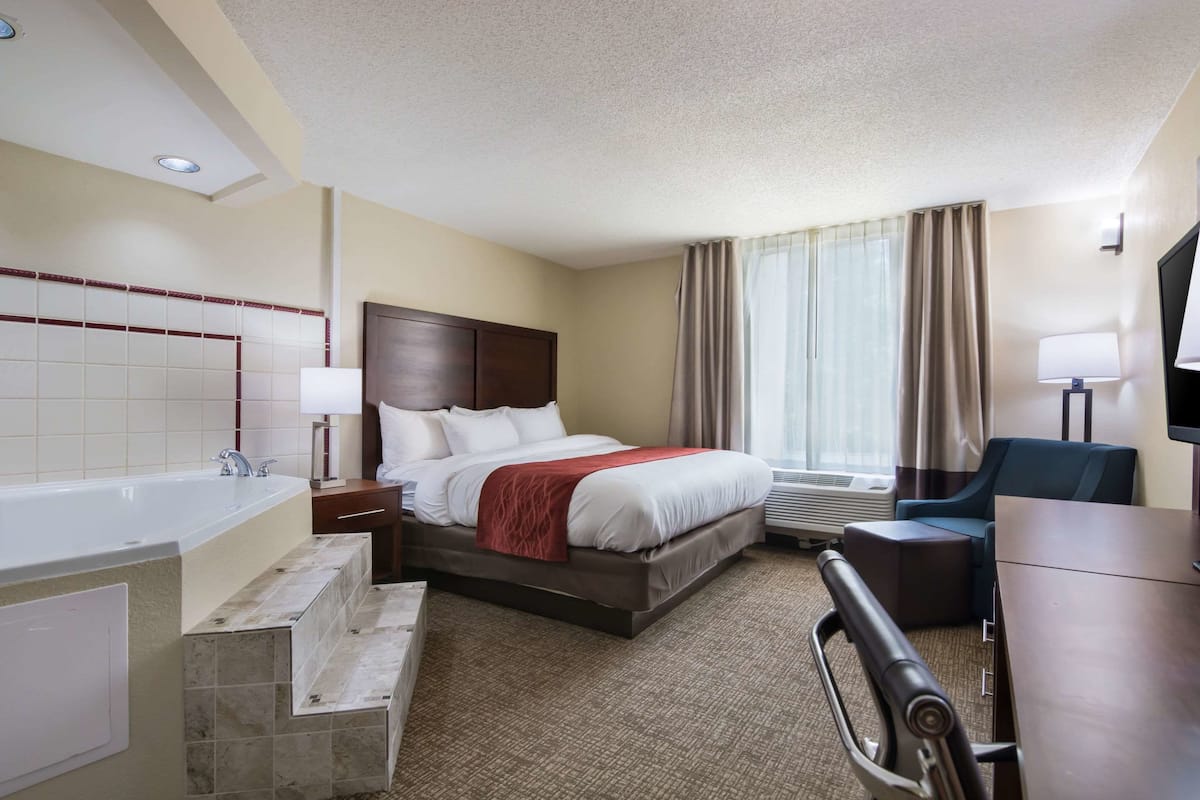 A hotel room with a bed, a desk, a chair, a TV, a window with curtains, and a bathroom with a bathtub and sink.