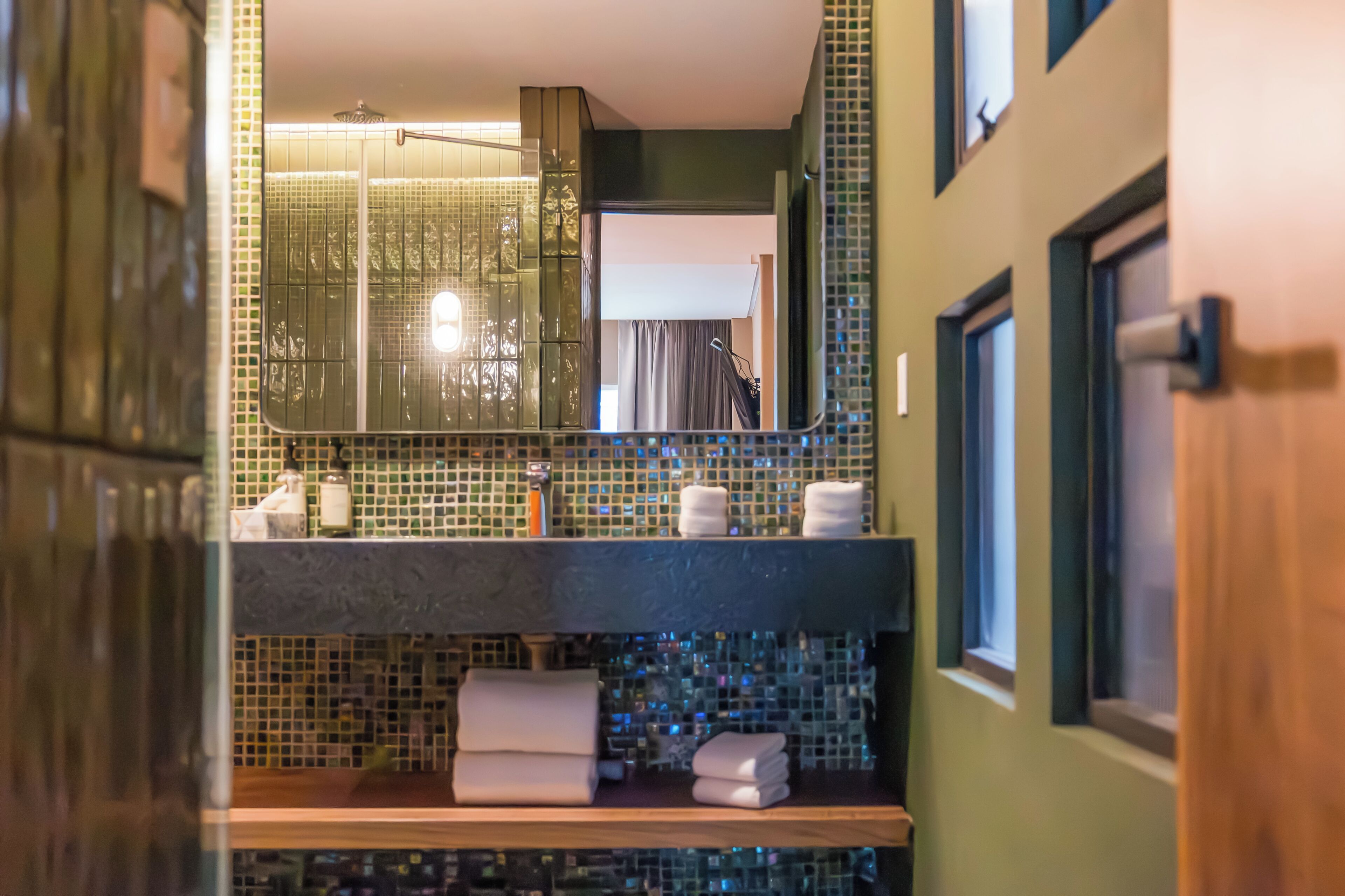 Un baño con pared de azulejos, un espejo, un lavamanos y una ventana.