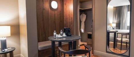 A hotel room with a round table, a chair, a bench, a robe, a mirror, and a coat rack.