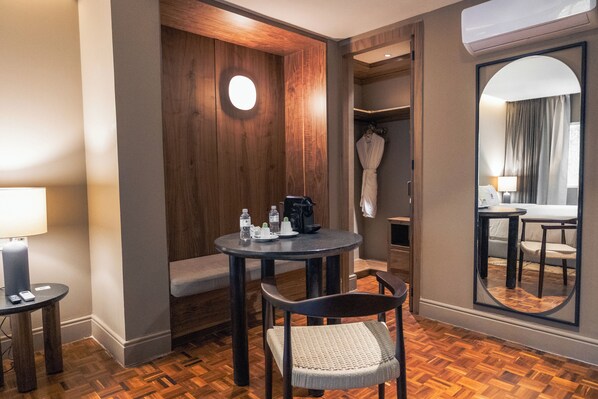 Una habitación de hotel con una mesa redonda, una silla, un banco, una bata, un espejo y un perchero.