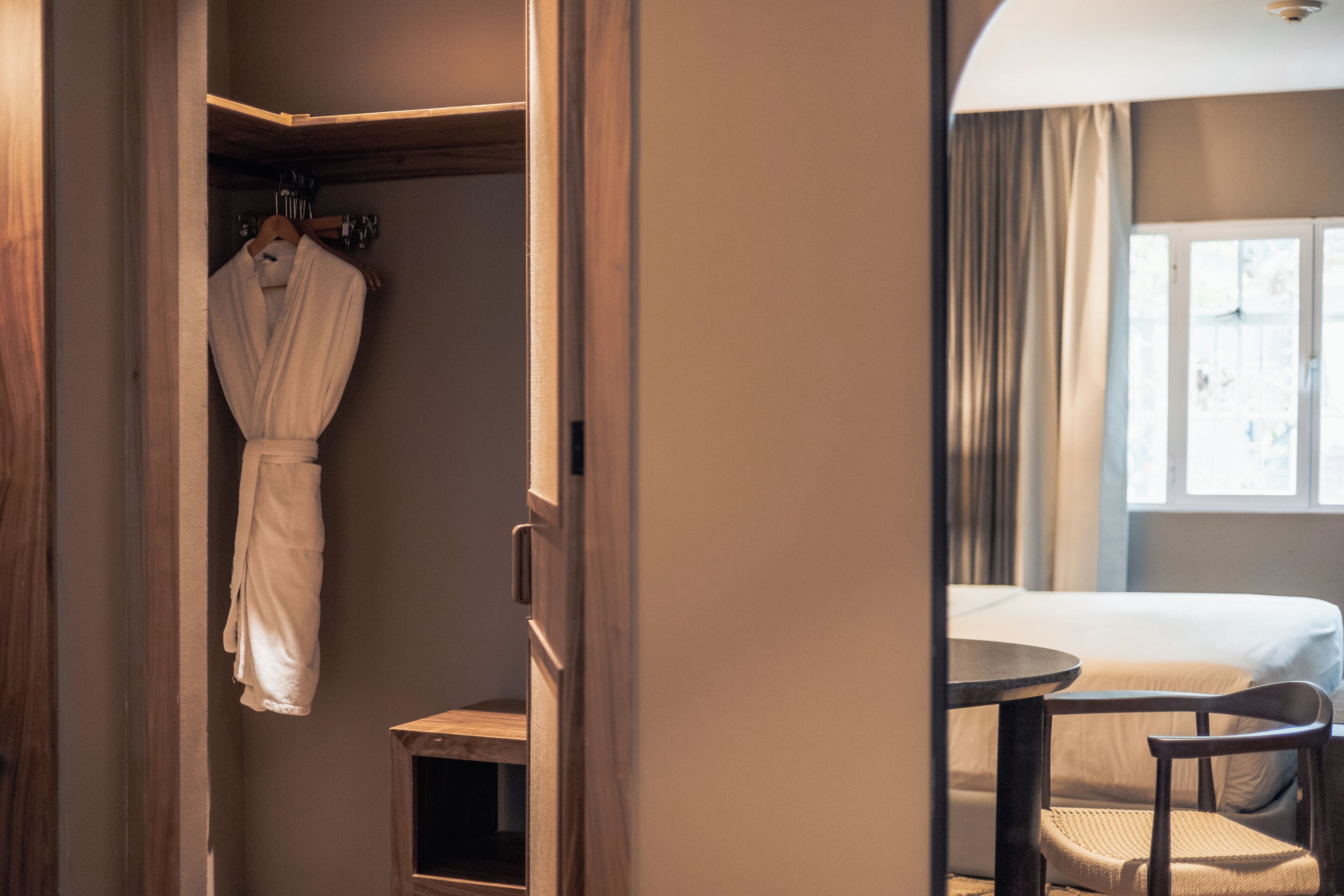 A hotel room with a bed, a small table, a chair, a robe hanging on a rack, and a window with curtains.