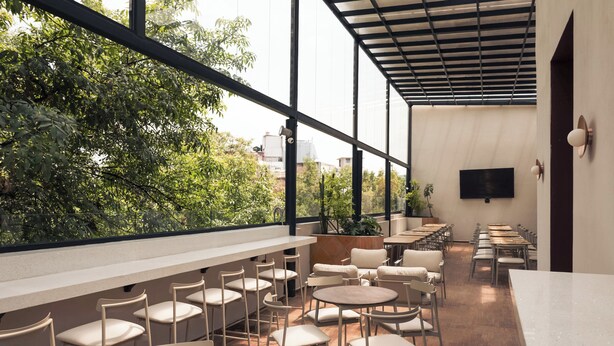 A modern indoor space with a long counter, multiple tables, and chairs, large windows with a view of greenery and buildings.
