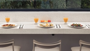 A breakfast table with plates of food and drinks, a window with a view of greenery, and a bar area with chairs.