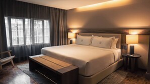 A hotel room with a large bed, bedside lamps, a wooden bench, and a view of the outdoors through the window.