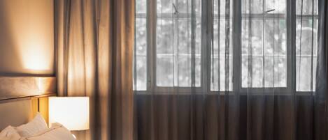 Una habitación de hotel con cama, mesita de noche, lámpara y ventana con cortinas.