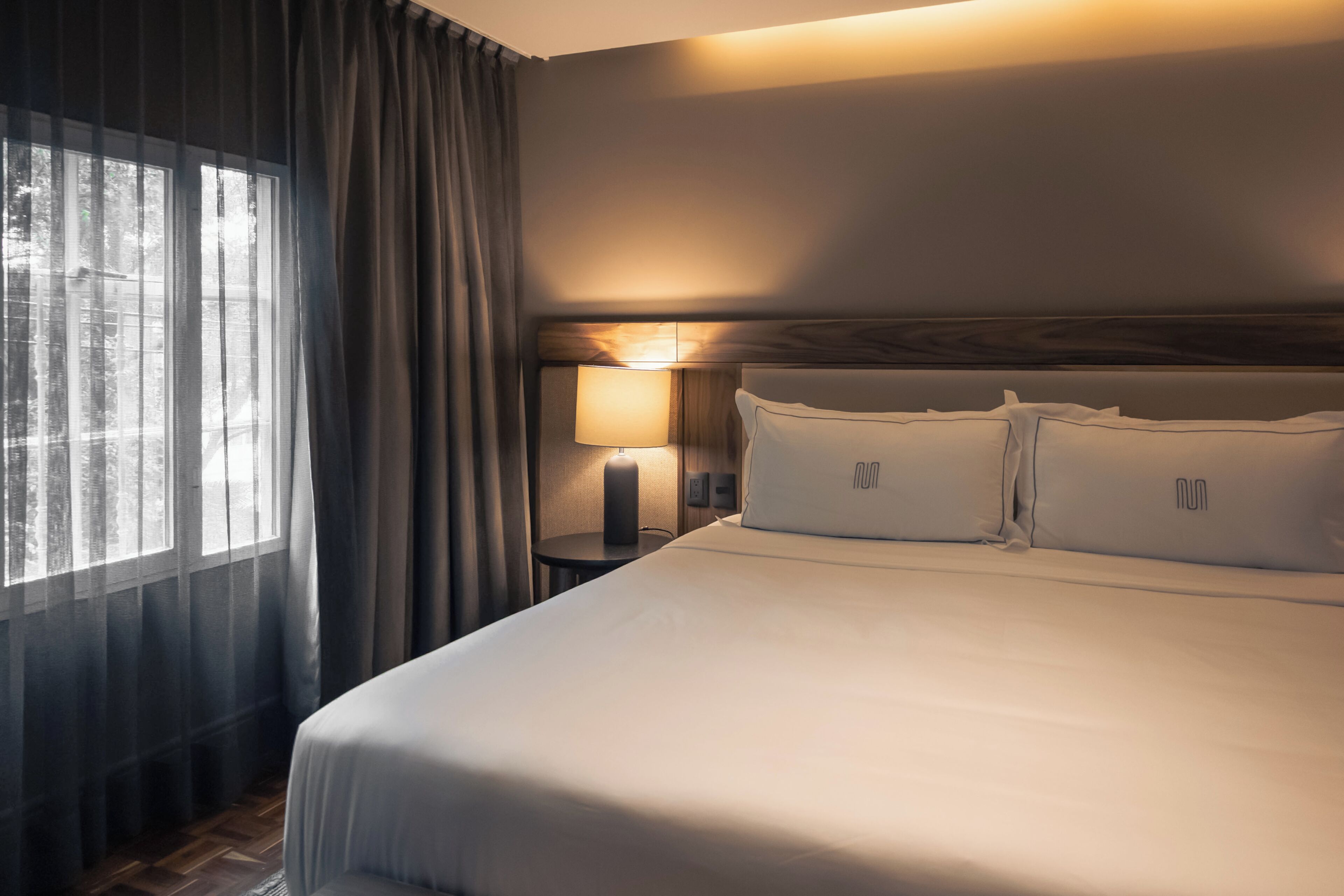 Habitación de hotel con una cama grande, una mesita de noche con lámpara y cortinas translúcidas en la ventana.